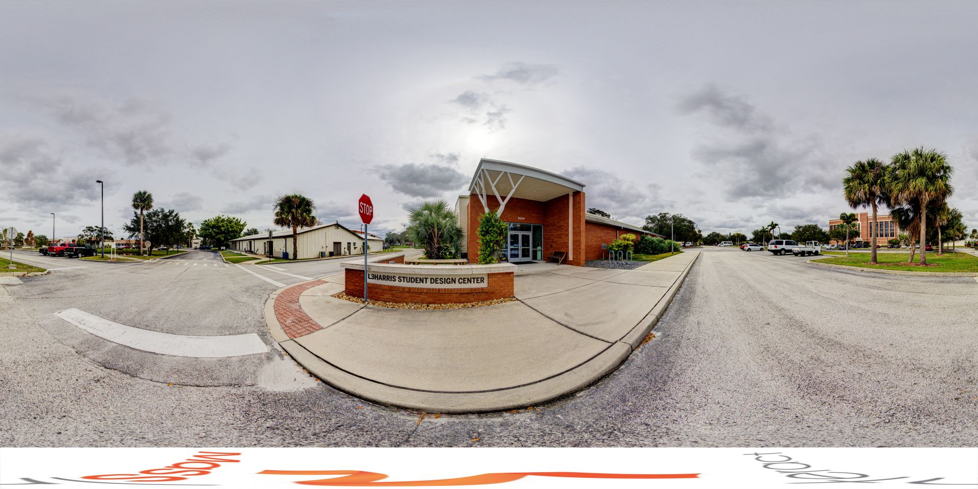 Panoramic view of L3Harris Student Design Center with a wide driveway, stop sign, and palm trees under an overcast sky